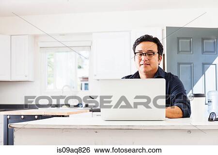 Stock Photograph - Mature businessman typing on laptop in kitchen. Fotosearch