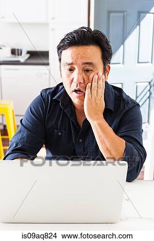 Mature businessman with hand on cheek working on laptop at kitchen table View Large Photo Image Picture - Mature businessman with hand on cheek working on laptop at kitchen table. Fotosearch