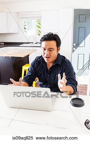 Stock Photography - Mature businessman with hands open working on laptop at kitchen table. Fotosearch