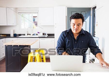 Stock Photo - Mature man typing on laptop at kitchen table. Fotosearch