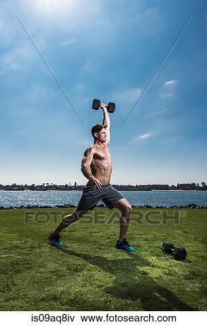 Stock Photo - Mid adult man lifting dumb bells at coast. Fotosearch