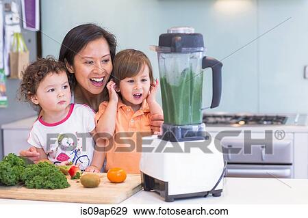 Stock Photo - Mid adult woman making smoothie for two toddlers in kitchen. Fotosearch