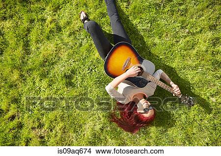 Picture - Overhead view of young woman lying on grass playing acoustic guitar. Fotosearch