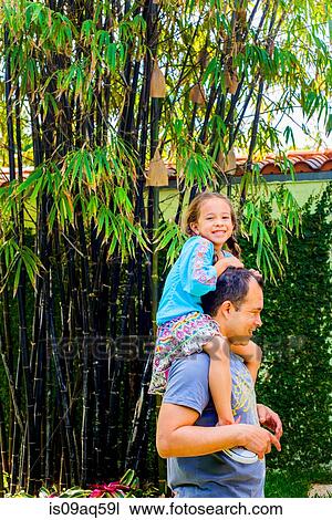 Stock Image - Portrait of girl getting a shoulder carry from father in garden. Fotosearch