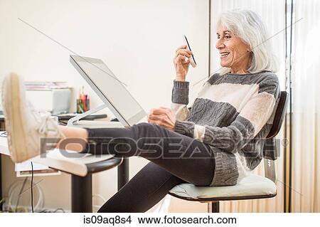 Picture - Portrait of senior woman sitting at computer holding smartphone with foot on desk. Fotosearch