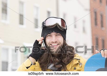 Stock Image - Portrait of young man holding snowboard and chatting on smartphone on street. Fotosearch