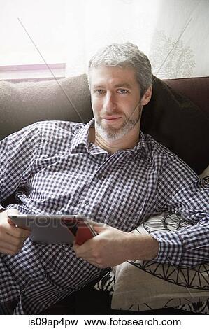 Stock Photo - Portrait of young man reclining on living room sofa using digital tablet. Fotosearch