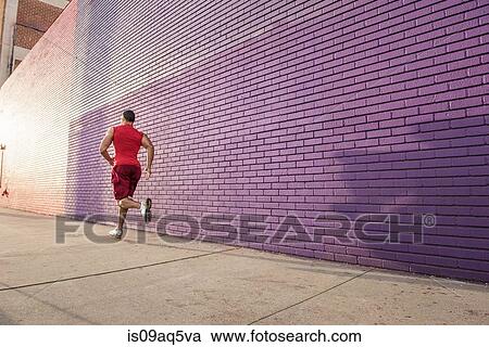 Stock Image - Rear view of male runner running along sidewalk. Fotosearch