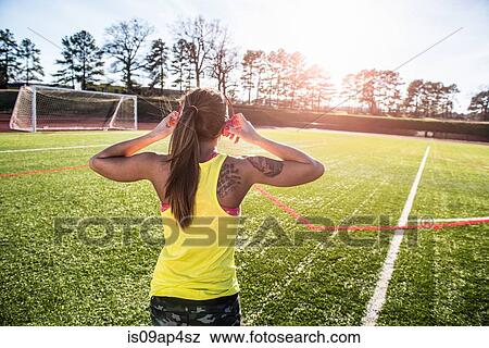 Stock Photo - Rear view of young female athlete putting on head phones on sports field. Fotosearch