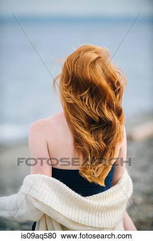 Stock Image - "Rear view of young woman with long red hair looking out to sea, Bainbridge Island, Washington State, USA". Fotosearch