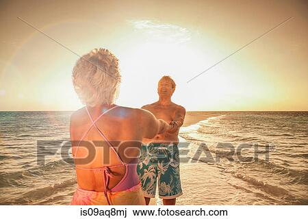 Stock Image - Senior couple holding hands on beach. Fotosearch