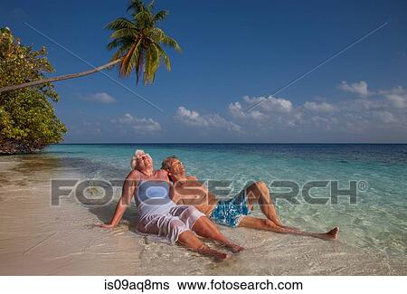 Stock Photograph - "Senior couple relaxing on beach, Maldives". Fotosearch