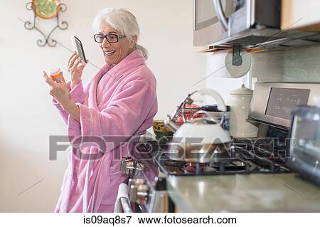 Stock Photo - Senior woman standing in kitchen holding prescription drugs bottle whilst talking on smartphone. Fotosearch