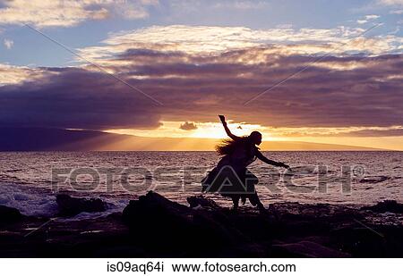 Stock Image - "Silhouetted woman hula dancing on coastal rocks wearing traditional costume at sunset, Maui, Hawaii, USA". Fotosearch