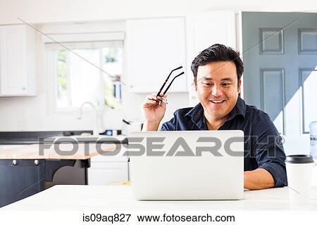 Stock Photo - Smiling mature businessman typing on laptop in kitchen. Fotosearch