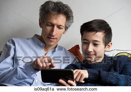 Stock Image - Teenage boy and father playing smartphone game on sofa. Fotosearch