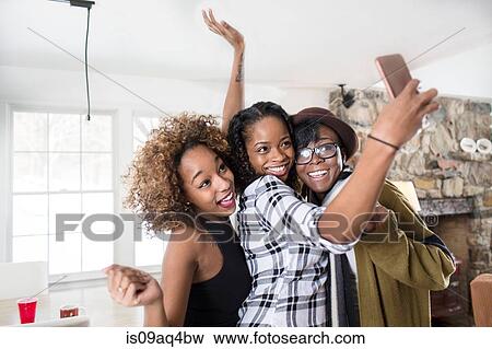 Stock Photo - Three young women posing for smartphone selfie in kitchen. Fotosearch