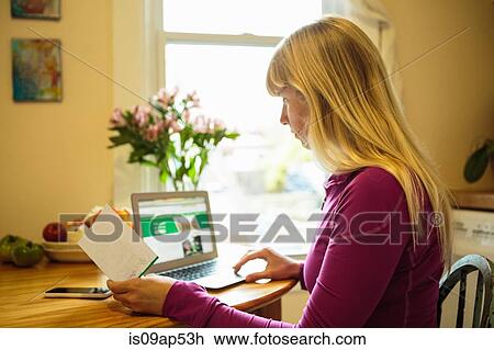 Stock Image - Woman working in kitchen table using laptop and reading instruction booklet. Fotosearch
