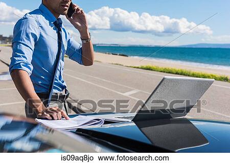 Picture - Young businessman at coast parking lot using laptop whilst talking on smartphone. Fotosearch