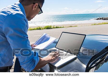 Young businessman at coast parking lot typing on laptop whilst reading paperwork View Large Photo Image Stock Photography - Young businessman at coast parking lot typing on laptop whilst reading paperwork. Fotosearch