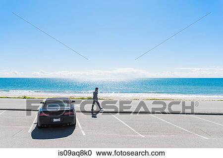 Stock Image - Young businessman at coastal parking lot walking whilst talking on smartphone. Fotosearch