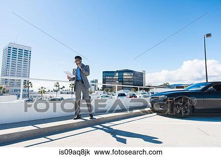 Stock Image - Young businessman on city rooftop car park chatting on smartphone. Fotosearch