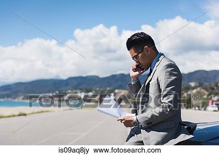 Young businessman sitting on car hood at coastal parking lot talking on smartphone View Large Photo Image Stock Photo - Young businessman sitting on car hood at coastal parking lot talking on smartphone. Fotosearch