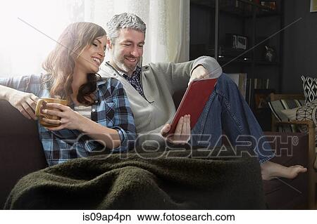 Young couple sitting on sofa drinking coffee and looking at digital tablet View Large Photo Image Picture - Young couple sitting on sofa drinking coffee and looking at digital tablet. Fotosearch