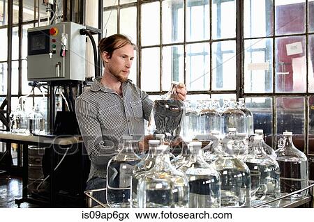 Stock Photo - Young male vodka distiller checking demijohn in distillery workshop. Fotosearch