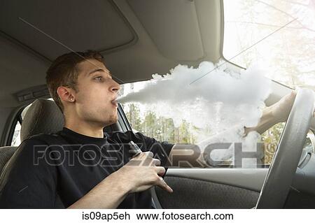 Stock Image - "Young man driving car, smoking ecigarette". Fotosearch