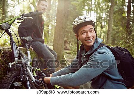 Stock Image - Young man fixing bike in forest while friend looks on. Fotosearch