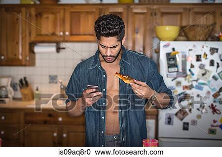 Stock Image - Young man holding cell phone and pizza in kitchen. Fotosearch
