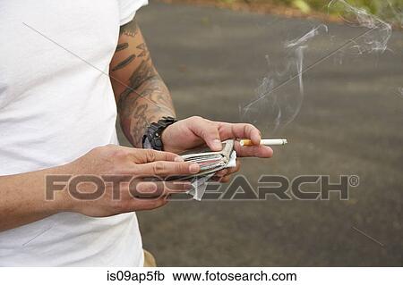 Stock Image - "Young man holding wallet and smoking cigarette, mid section". Fotosearch