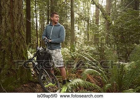 Stock Image - "Young man in forest, leaning mountain bike against tree". Fotosearch