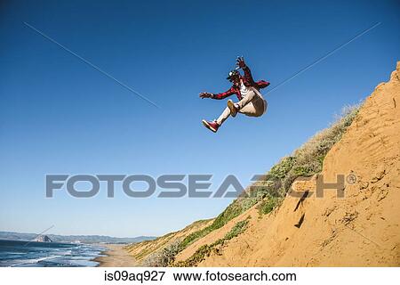 Stock Photo - "Young man jumping down sandy hill, in mid air". Fotosearch