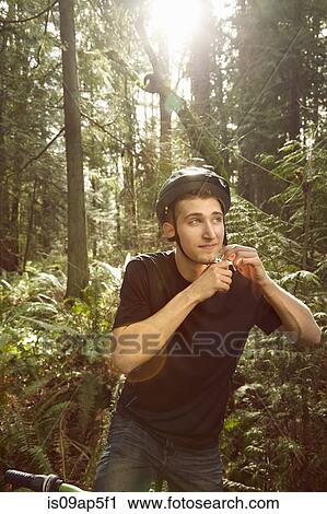 Stock Image - "Young man on bike in forest, securing bicycle helmet". Fotosearch