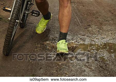 Picture - "Young man pushing bike on dirt track, low section". Fotosearch
