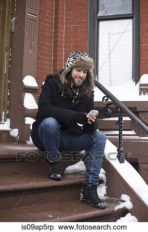 Stock Photography - Young man reading text on smartphone on snow covered front door step. Fotosearch
