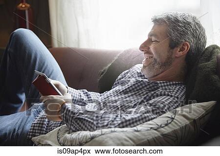 Young man reclining on living room sofa using digital tablet View Large Photo Image Stock Photo - Young man reclining on living room sofa using digital tablet. Fotosearch