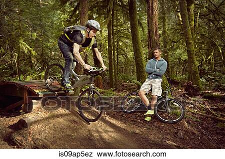 "Young man riding mountain bike over ramp, in forest, while friend watches" View Large Photo Image Stock Photo - "Young man riding mountain bike over ramp, in forest, while friend watches". Fotosearch