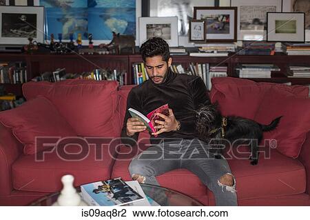 Stock Image - Young man sitting on sofa reading book. Fotosearch