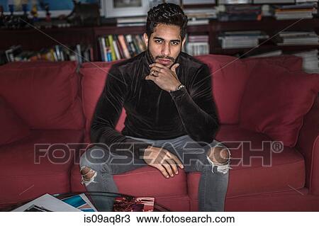 Stock Image - Young man sitting on sofa with hand on chin. Fotosearch