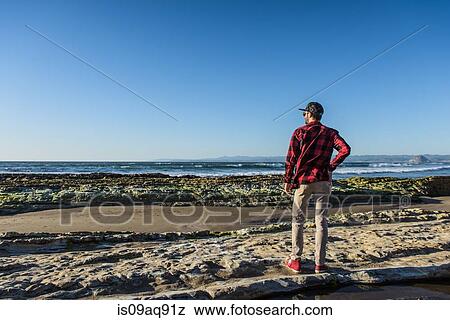 Stock Photo - "Young man standing on beach looking at ocean, rear view". Fotosearch