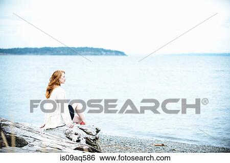 Stock Photograph - "Young woman sitting on beach looking out to sea, Bainbridge Island, Washington State, USA". Fotosearch