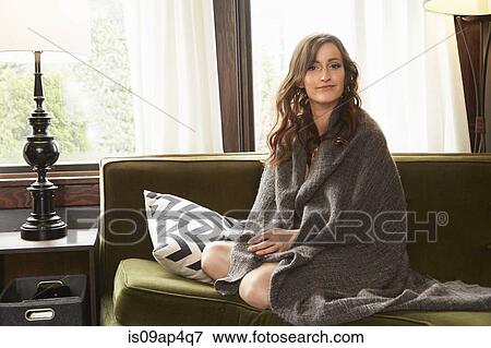 Stock Photo - Young woman sitting on living room sofa wrapped in blanket. Fotosearch