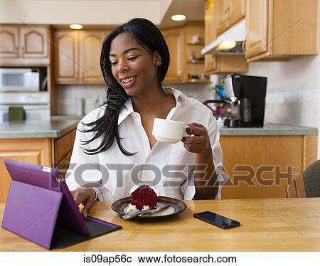 Stock Image - Young woman using digital tablet at kitchen table. Fotosearch