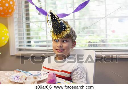 Boy in party hat sitting at table eating birthday cake pulling funny face View Large Photo Image Stock Photograph - Boy in party hat sitting at table eating birthday cake pulling funny face. Fotosearch