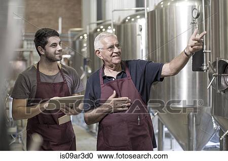 Stock Image - Brewers in brewery standing next to stainless steel tanks. Fotosearch