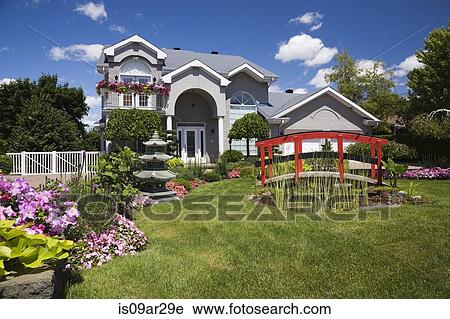 Stock Image - "Grey brick cottage style residential home and landscaped manicured garden with red footbridge, Quebec, Canada". Fotosearch