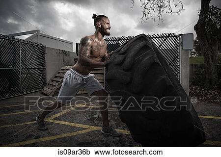 Stock Image - Male boxer with gritted teeth pushing truck tyre in yard. Fotosearch